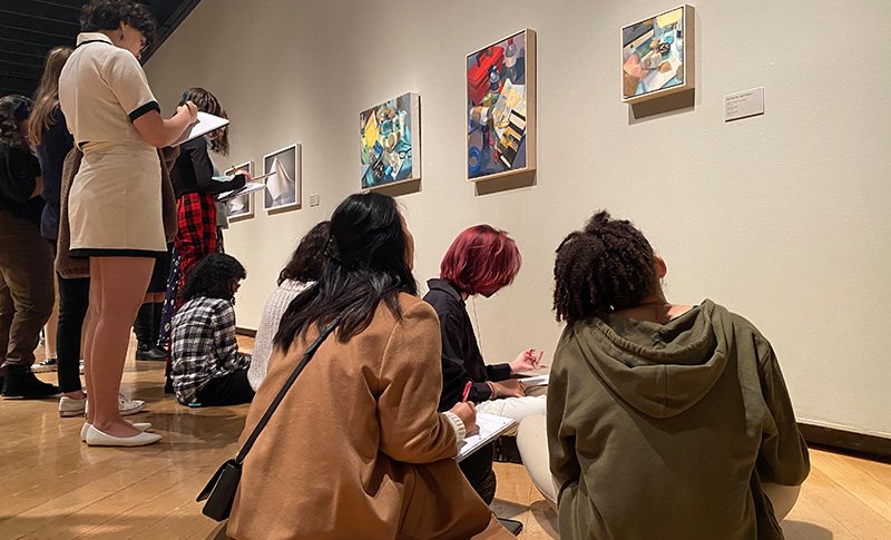 Several students sketching on note pads, facing a wall of paintings.