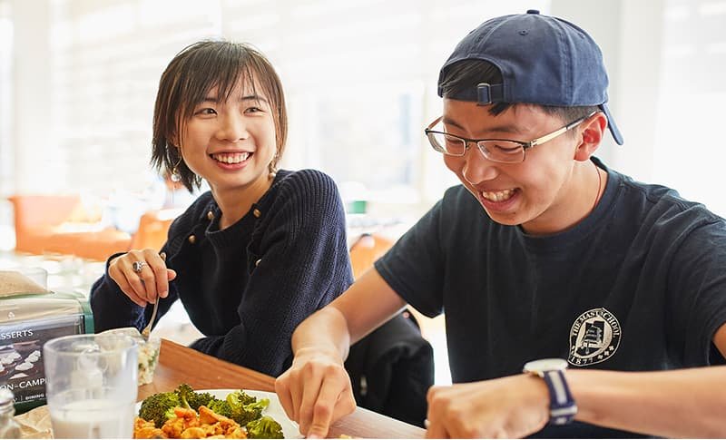 Two students smiling and eating together in a sunny dining hall.