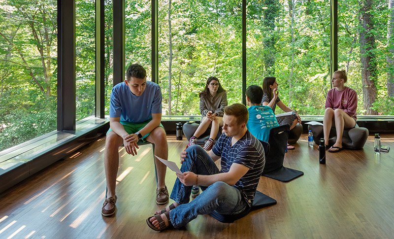 Several students sitting peacefully in chairs and meditation seats in a  room overlooking a green wooded area.